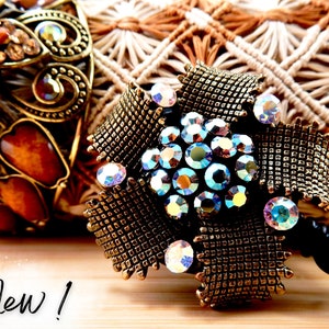 May include: A gold-toned metal flower-shaped badge reel with a large cluster of iridescent rhinestones in the centre. The reel is attached to a black retractable cord. The word "New!" is written on a wooden surface in the foreground.