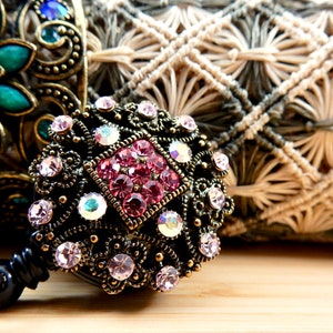 Pu&ograve; includere: Primo piano di un portabadge decorato con strass rosa e trasparenti. Il portabadge &egrave; in metallo e ha una clip nera.