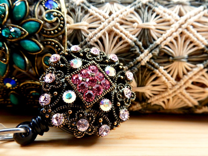 Pu&ograve; includere: Primo piano di un portabadge decorato con strass rosa e trasparenti. Il portabadge &egrave; in metallo e ha una clip nera.