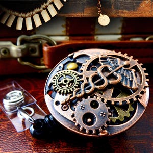 May include: A steampunk-style badge reel with a bronze and brass gear design, featuring a Caduceus symbol. The reel is set against a dark brown leather background, with a clear plastic holder and a black retractable cord.