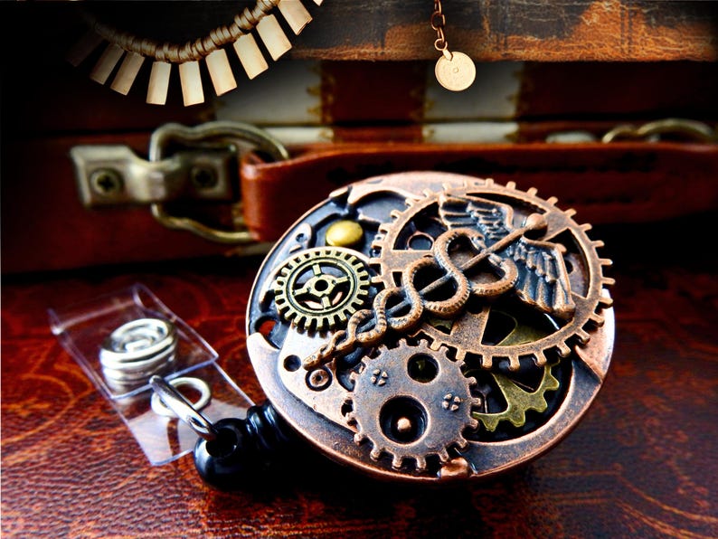 May include: A steampunk-style badge reel with a bronze and brass gear design, featuring a Caduceus symbol. The reel is set against a dark brown leather background, with a clear plastic holder and a black retractable cord.