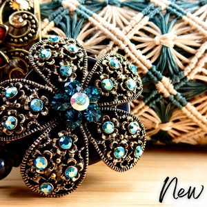 May include: A teal blue jeweled flower charm with a gold filigree design. The charm is attached to a black retractable badge holder. The charm is sitting on a wooden surface.