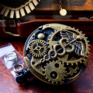 Puede incluir: Un portatarjetas de estilo steampunk con intrincados engranajes de lat&oacute;n y un s&iacute;mbolo m&eacute;dico del caduceo. El portatarjetas tiene una base negra y un cord&oacute;n retr&aacute;ctil. Se ve una peque&ntilde;a bolsa de accesorios plateados.