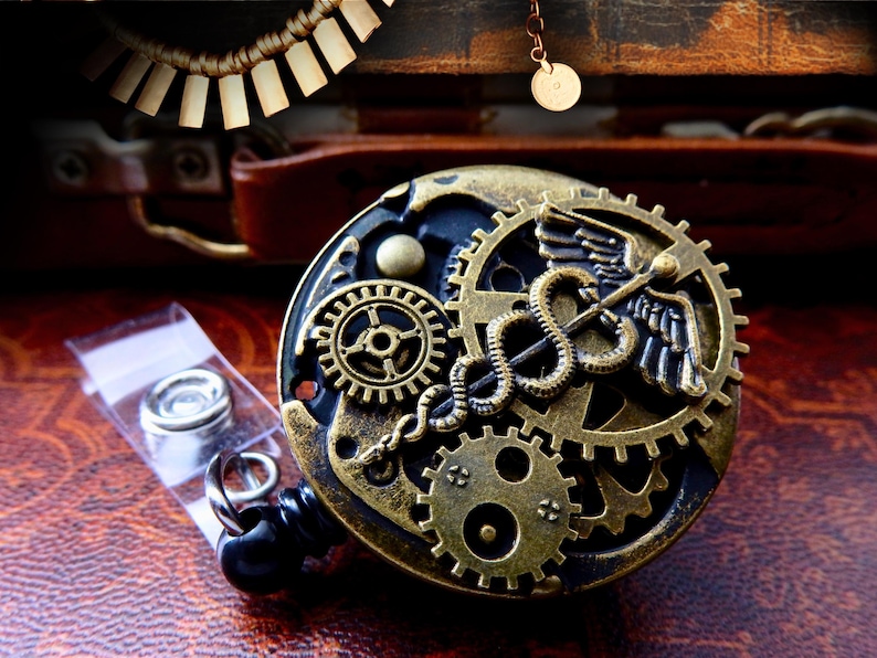Puede incluir: Un portatarjetas de estilo steampunk con intrincados engranajes de lat&oacute;n y un s&iacute;mbolo m&eacute;dico del caduceo. El portatarjetas tiene una base negra y un cord&oacute;n retr&aacute;ctil. Se ve una peque&ntilde;a bolsa de accesorios plateados.