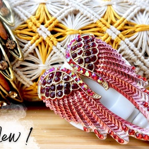 May include: A pair of red and pink angel wings with gold accents and rhinestones. The wings are attached to a white clip.