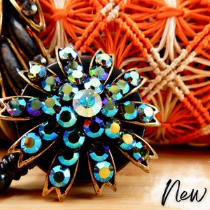 May include: A black retractable badge holder with a decorative flower-shaped charm. The charm is made of gold-toned metal and features iridescent blue and green rhinestones.