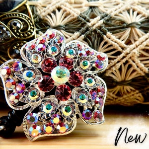May include: A silver flower-shaped badge reel with a large red rhinestone centre surrounded by smaller rhinestones in various colours, including pink, green, and purple. The reel is attached to a black retractable cord.
