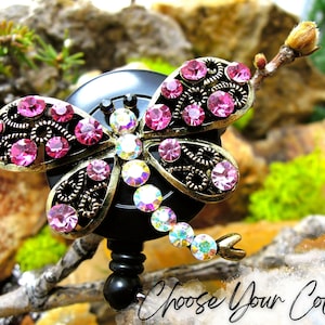 May include: A black retractable badge holder with a gold dragonfly design. The dragonfly is decorated with pink and clear rhinestones. "Choose Your Color" is written in cursive at the bottom of the image.