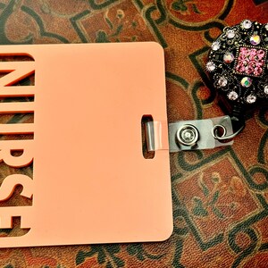 Pu&ograve; includere: Un porta badge in plastica color pesca con la parola "NURSE" ritagliata sul lato sinistro. Il porta badge ha un cinturino in plastica trasparente con un fermaglio metallico e un rotolo retrattile per badge nero con un design di strass rosa e nero.