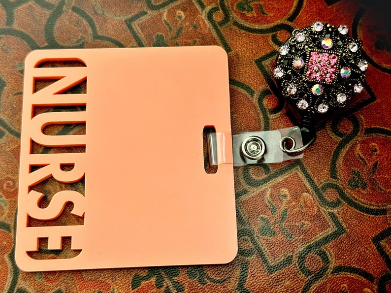 Pu&ograve; includere: Un porta badge in plastica color pesca con la parola "NURSE" ritagliata sul lato sinistro. Il porta badge ha un cinturino in plastica trasparente con un fermaglio metallico e un rotolo retrattile per badge nero con un design di strass rosa e nero.