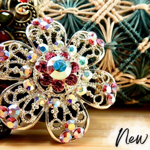 May include: A silver flower-shaped brooch with a large, multi-faceted, pink and blue rhinestone centre. The brooch is surrounded by smaller rhinestones and has a delicate, intricate design.