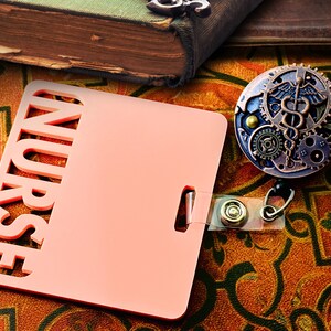 May include: A pink badge holder with the word "NURSE" cut out, next to a steampunk-style badge reel with gears and a medical symbol. The items rest on a patterned surface.