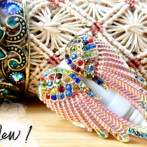 Może przedstawiać: Para różowych skrzydeł anielskich ozdobionych tęczowymi kryształkami. Skrzydła są wykonane z metalu i mają delikatny, kobiecy design.