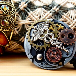 May include: A silver and bronze steampunk-style badge reel with a medical caduceus symbol and gears. The reel is attached to a black retractable cord.