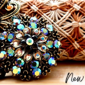 May include: A gold-toned brooch with a floral design featuring iridescent blue and black rhinestones. The brooch is set against a woven fabric background.