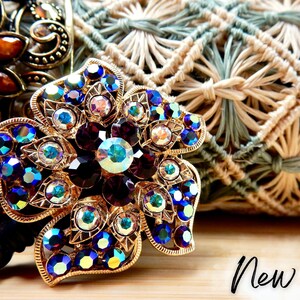 May include: A gold-tone flower-shaped brooch with purple and iridescent blue rhinestones. The brooch is on a woven background.