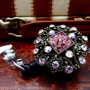 Pu&ograve; includere: Un porta badge retrattile nero con un decoro decorativo, ornato, dorato, con strass rosa e trasparenti.