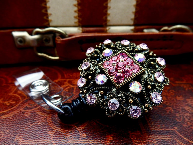 Pu&ograve; includere: Un porta badge retrattile nero con un decoro decorativo, ornato, dorato, con strass rosa e trasparenti.