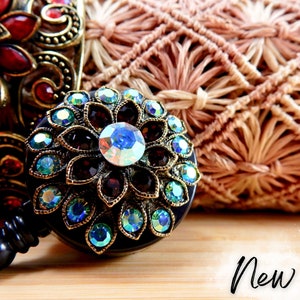 May include: A black retractable badge holder with a round, ornate, jeweled design. The design features a central blue iridescent jewel surrounded by smaller, burgundy jewels and iridescent green jewels.