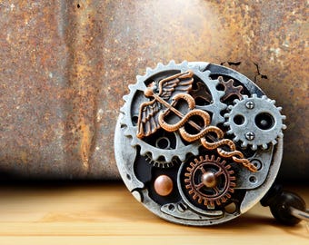 May include: A steampunk-style badge featuring a medical caduceus symbol with wings and a snake, set against a backdrop of interlocking gears in silver, copper, and black. The badge is on a wooden surface.
