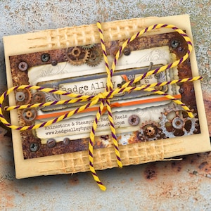 May include: A rectangular package wrapped in beige paper, tied with a yellow and purple striped cord. The package features a decorative card with gears, rivets, and the text "Badge Alley". The background is a textured, rusty surface.