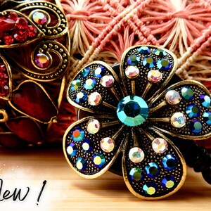 May include: A gold-toned retractable badge holder featuring a blue and iridescent flower design. The flower has a large teal centre stone and is surrounded by smaller blue and clear rhinestones.