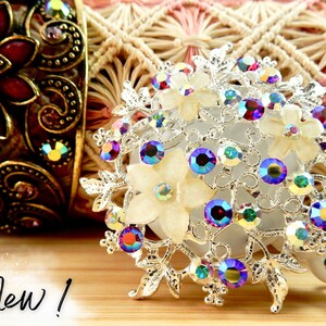 May include: A silver-toned, ornate, floral-shaped badge holder with iridescent rhinestones and white flowers. The badge holder is attached to a retractable cord. "New!" is written on the wooden surface below the badge holder.