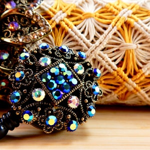 May include: A close-up of a decorative metal charm with blue and clear iridescent rhinestones. The charm is attached to a black cord and is resting on a woven surface.