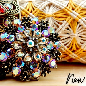 May include: Ornate, antique-style brooch with iridescent, multi-colored rhinestones set in a bronze-toned metal. The brooch has a floral design and the word "New!" in a handwritten font.