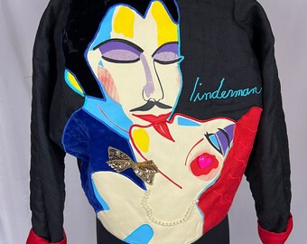 May include: Black jacket with a colorful abstract design of two faces kissing. The design includes yellow, blue, red, and purple accents, with the name "linderman" embroidered in blue. The jacket has red cuffs and a black velvet collar.
