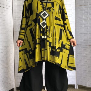 May include: A long-sleeved tunic featuring a cowl neckline and a bold olive green and black abstract brushstroke pattern. The garment is paired with solid black wide-leg trousers and a long black cord necklace with three geometric diamond-shaped pendants.