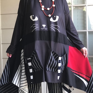 May include: Black tunic-style top with a cat face design, long sleeves, and asymmetrical hem. Features pockets with vertical stripes and red accents. Worn with black and white striped pants and a red necklace.