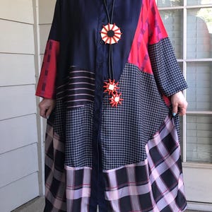 May include: A long, button-down dress in navy blue, red, and black patchwork. The dress features a variety of patterns, including plaid, stripes, and a grid design. A long necklace with a sunburst pendant is worn with the dress. The dress has long sleeves.