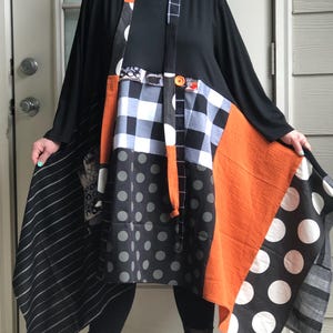 May include: A long-sleeved black tunic dress featuring a patchwork design with various patterns including black and white buffalo check, large white polka dots on black, grey polka dots, thin white stripes, and a solid burnt orange fabric panel.