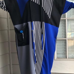 May include: A color-blocked dress in black, blue, gray, and white. The dress features a large pocket with a blue button and a striped pattern on the sleeves and front. The dress has a loose, flowing design.