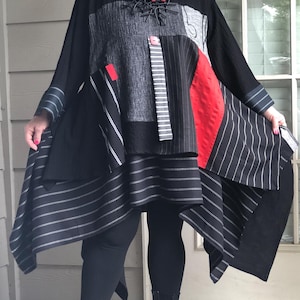 May include: A long-sleeved tunic featuring a patchwork design of black, grey, and red fabrics. The garment incorporates various textures, including pinstriped panels, solid black sections, and a bold red accent. It has an asymmetrical hemline.