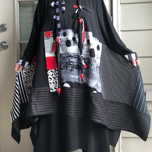 May include: A long-sleeved tunic dress featuring a black base with a patchwork design of white polka dots, thin white stripes, and a textured gray and black abstract print. The garment includes a red button, red beads, and fabric ties.