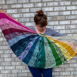 以下が含まれることがあります： ピンク、パープル、ブルー、グリーン、イエローなど、さまざまな色を使用したカラフルなストライプのニットショールを着用した人。ショールは人の肩にかけられ、手で持たれています。