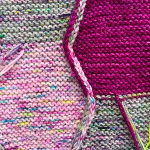 May include: Close-up of a knitted fabric with different colored squares. The squares are pink, purple, gray, and green. The fabric is made of yarn and has a textured surface.