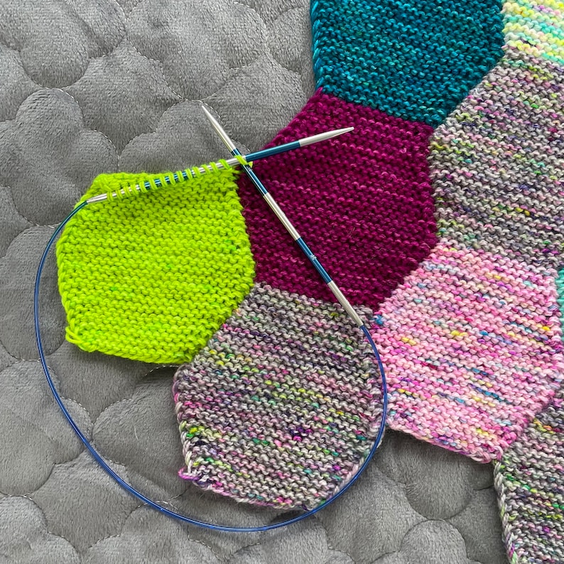 May include: A close-up of a colorful knitted blanket with different colored squares. The blanket is being worked on with knitting needles.