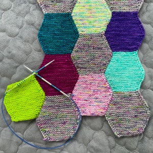 May include: A close-up of a colorful knitted blanket with a variety of hexagonal shapes in shades of green, purple, pink, and gray. Two knitting needles are visible in the image.