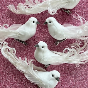 May include: Several white decorative bird clips with gray beaks and feathery tails are arranged on a pink glitter surface. The birds have a soft, textured appearance and are designed to be clipped onto various items.