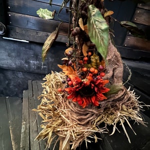 May include: A fall-themed centerpiece with a burlap base, dried leaves, and a large orange sunflower. The centerpiece is decorated with small red berries and is sitting on a dark wooden surface.