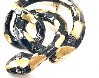 May include: A coiled snake brooch in black, gold, and silver tones. The brooch features a detailed design with a textured appearance. The snake's head is visible, and the brooch has a pin closure on the back. The jewelry piece is set against a white background.