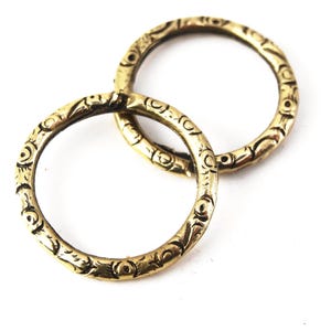 May include: Two circular metal rings with a dull gold finish, each featuring a repeating embossed pattern of small circles and curved lines along the entire outer surface. The rings are positioned overlapping each other against a plain white background.