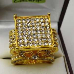 May include: A gold ring with a large square top, set with many clear, sparkling stones. The ring has a wide band also decorated with smaller stones. It is displayed in a white and brown box.