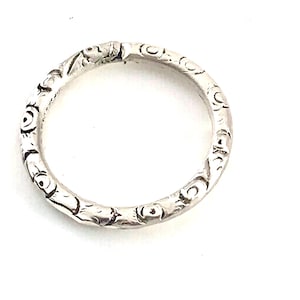 May include: A silver-toned circular ring featuring a textured surface with repeating embossed circular and linear patterns. The metal band has a slightly uneven, hand-crafted appearance with a metallic finish and a visible seam at the top.