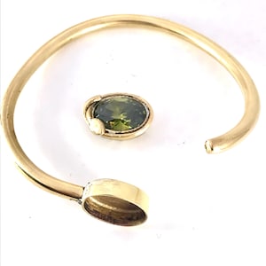 May include: A gold-coloured cuff bracelet featuring an oval, olive-green gemstone. The bracelet has a simple, open design with a gemstone setting on one end. The gemstone is surrounded by gold-coloured metal.