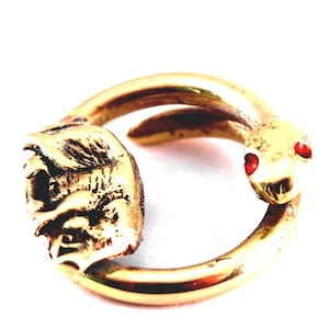 Puede incluir: Un anillo dorado con diseño de serpiente. La cabeza de la serpiente tiene ojos rojos y está opuesta a una cabeza de león detallada. El anillo está sobre un fondo blanco.