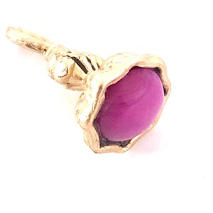 May include: A gold-colored jewelry piece featuring a vibrant magenta-colored stone. The stone is oval-shaped and set within a textured, organic-looking gold setting. The piece has a stem-like extension.